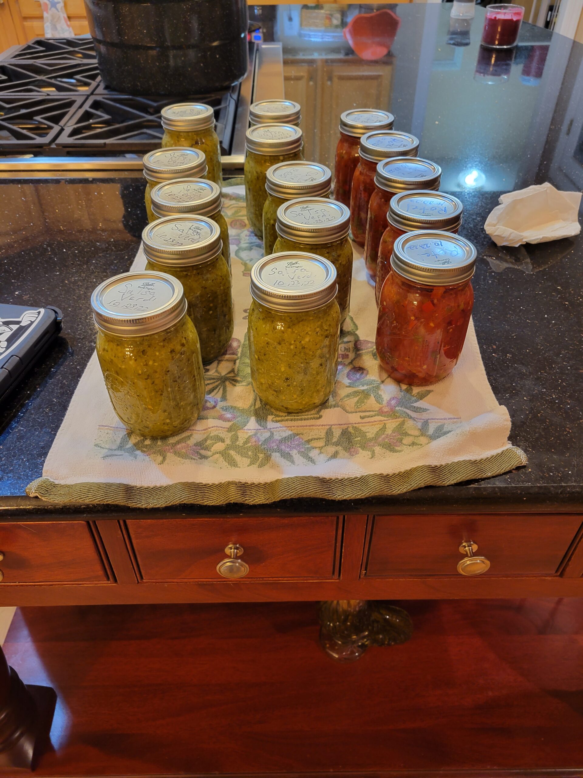 Douglas Davis - 20221024_072525 Homemade canned jars of green salsa and red salsa displayed on a kitchen counter, with a black pot and other kitchen items in the background. The jars are labeled with dates, showcasing a successful canning session.