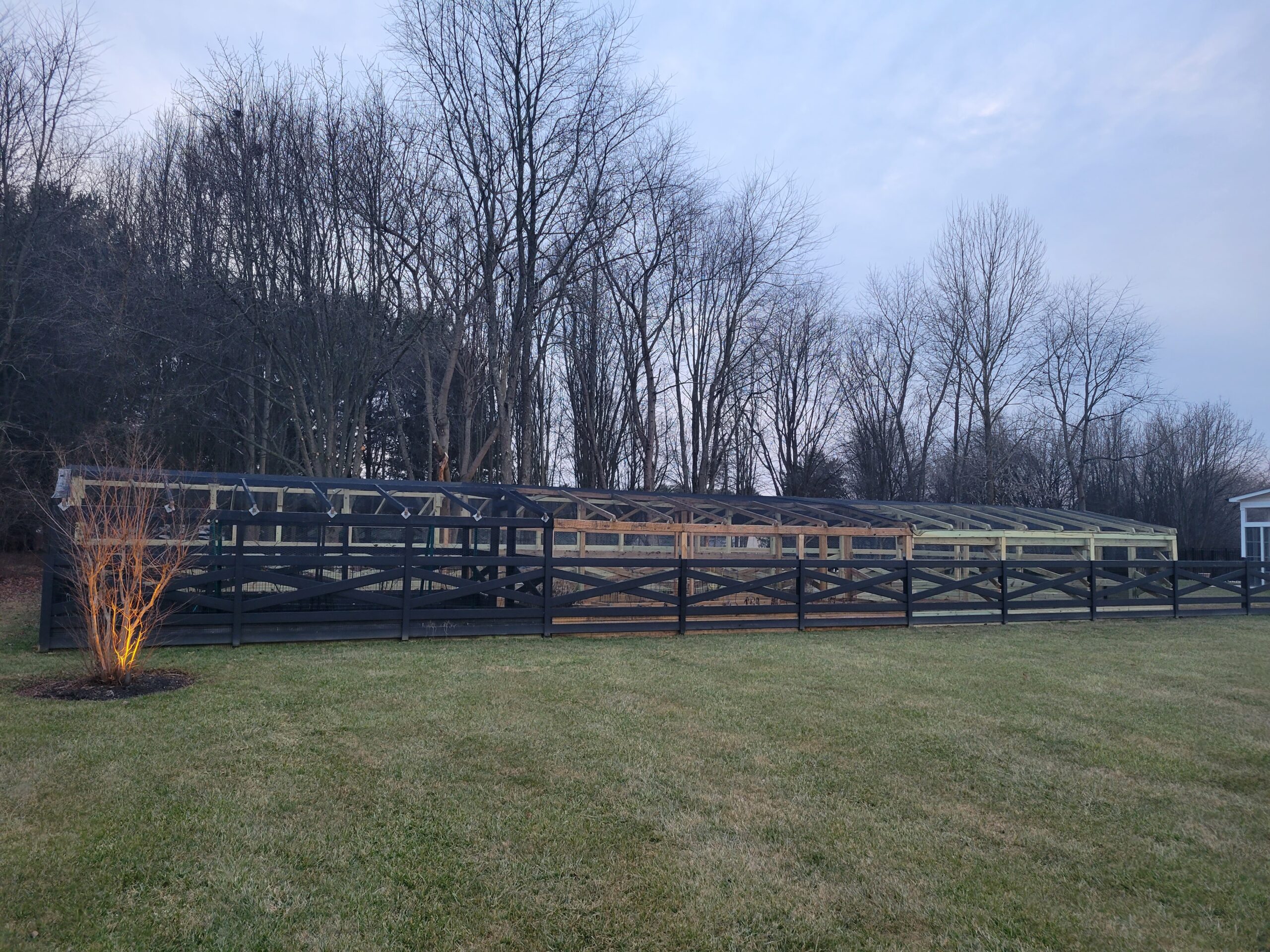 Greenhouse structure surrounded by a black wooden fence, set against a backdrop of bare trees and an overcast sky, with grassy lawn in the foreground.