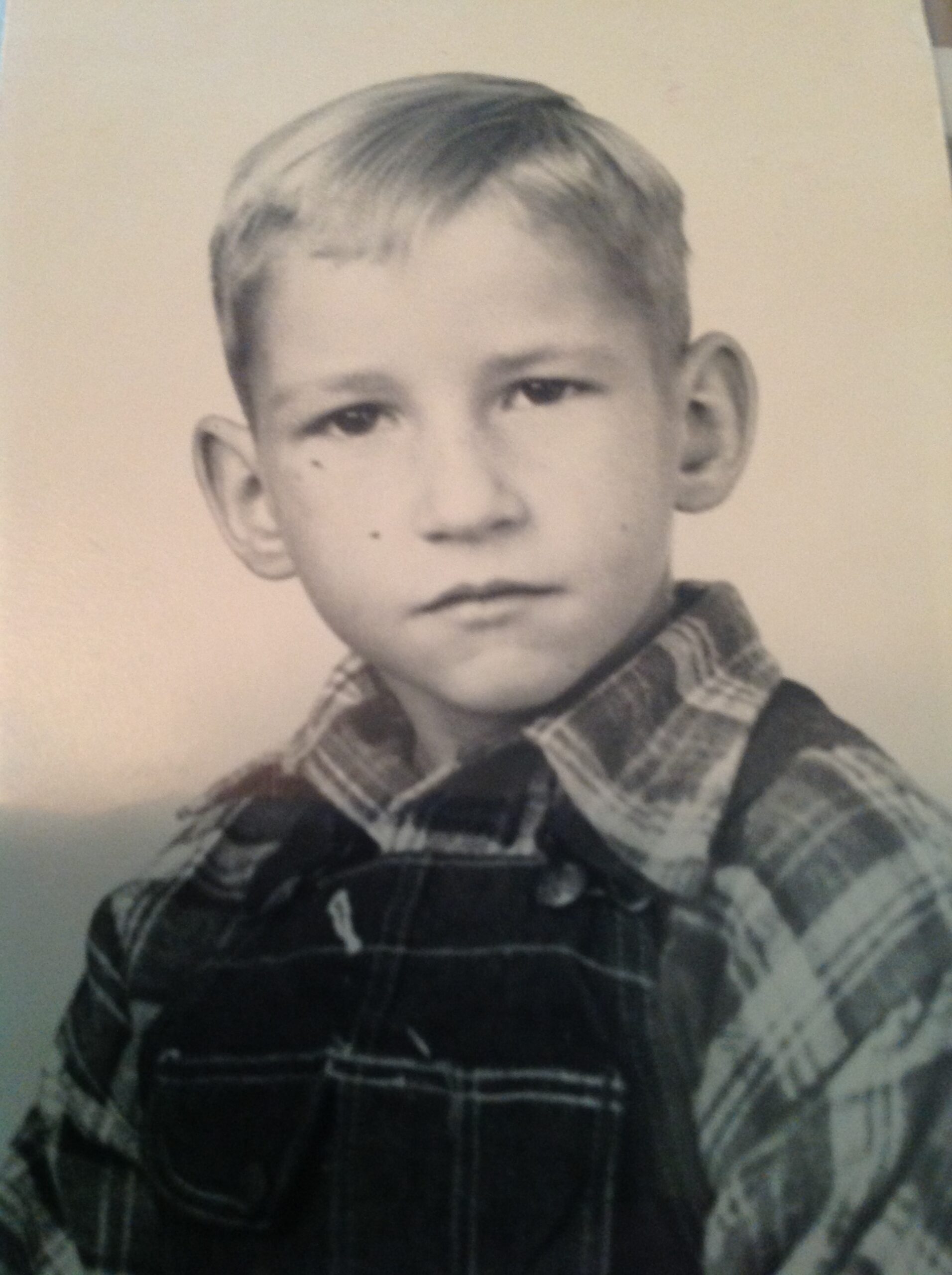 Black and white portrait of a young boy with short hair, wearing a plaid shirt and overalls, looking directly at the camera with a serious expression.