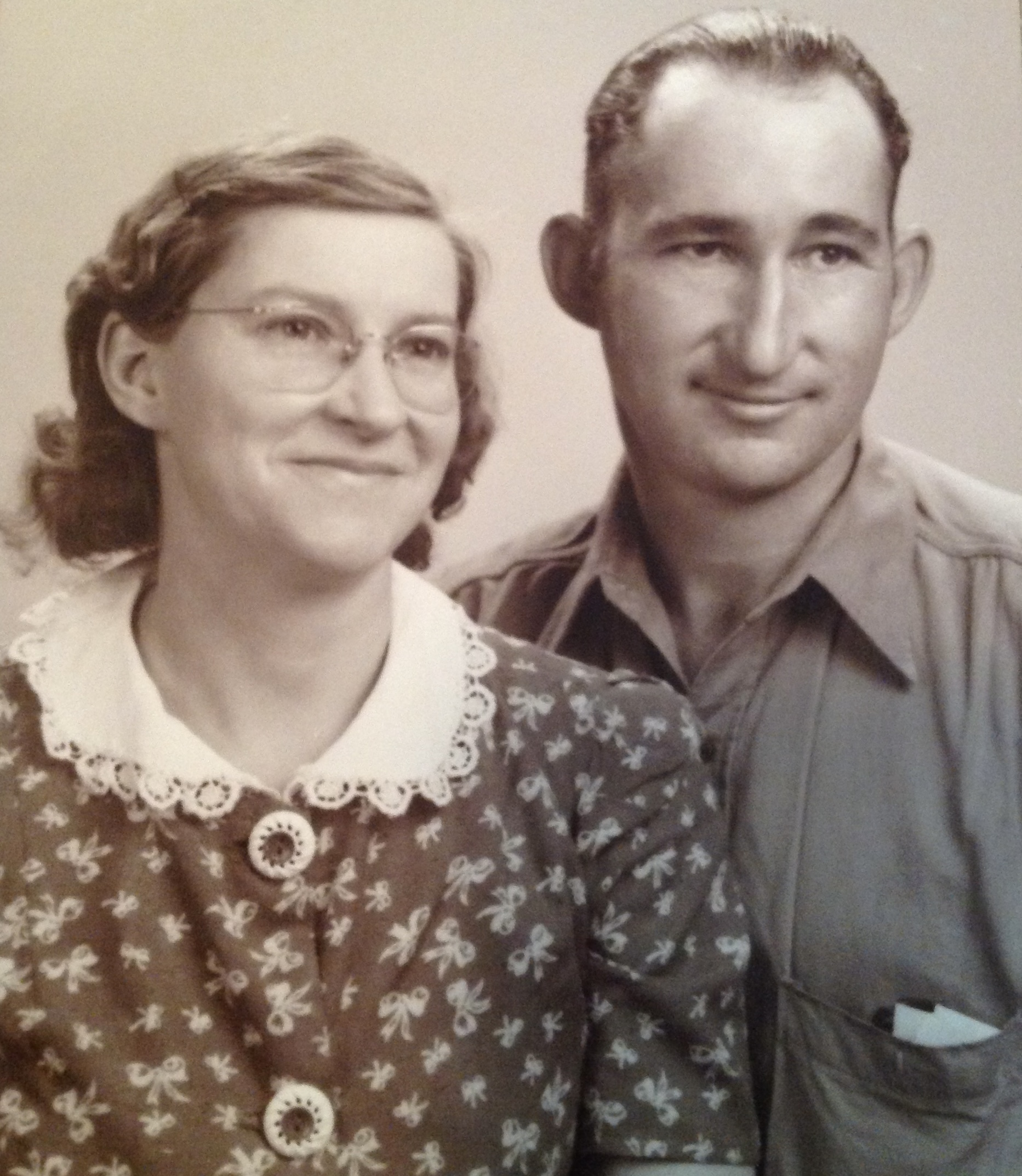 Black and white portrait of a smiling couple, featuring a woman in a floral dress with a lace collar and glasses, alongside a man in a collared shirt, both posing closely together.
