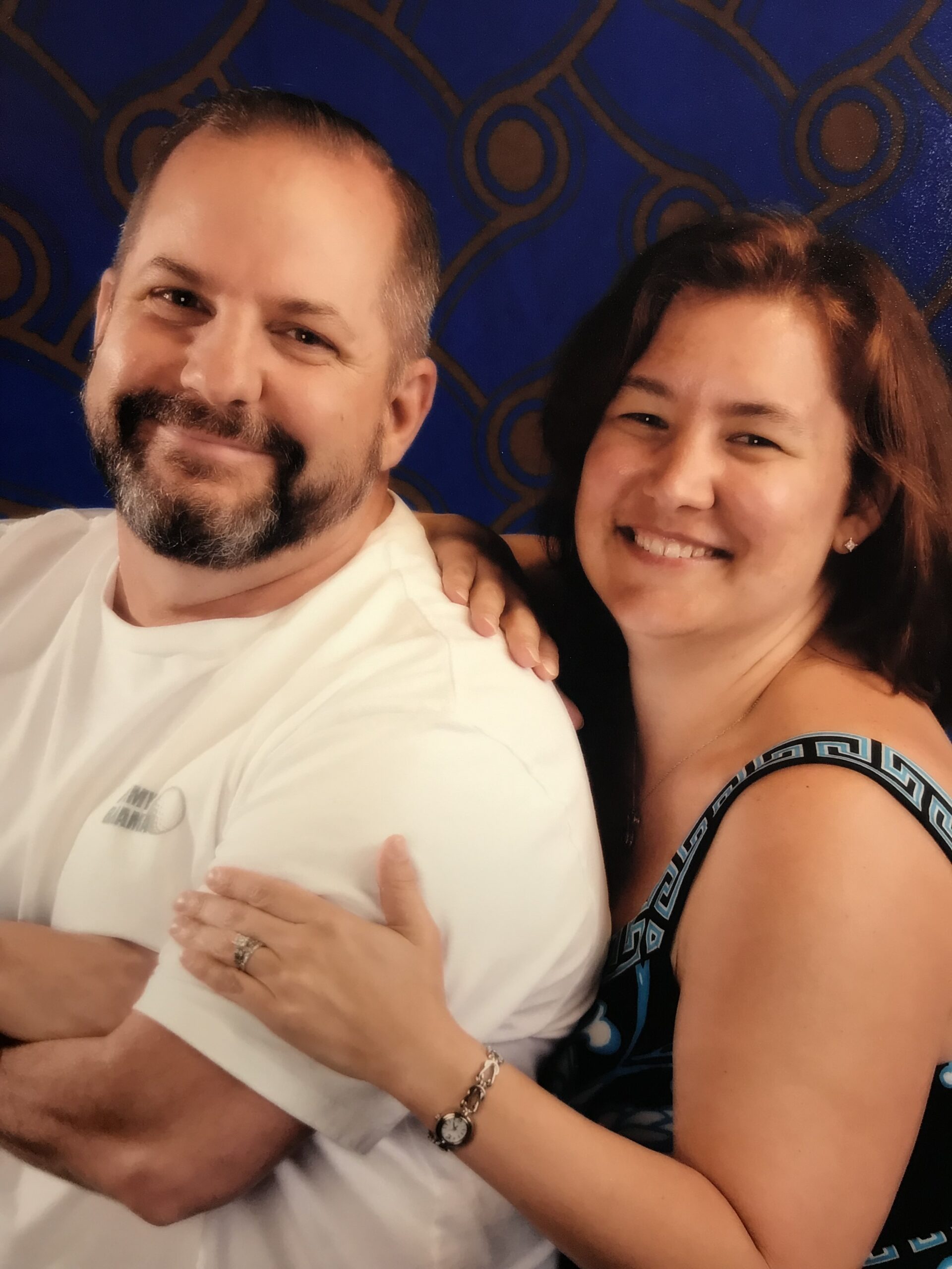 Smiling couple posing together, with the man wearing a white t-shirt and the woman in a patterned dress, showcasing a warm and affectionate moment against a blue and gold background.
