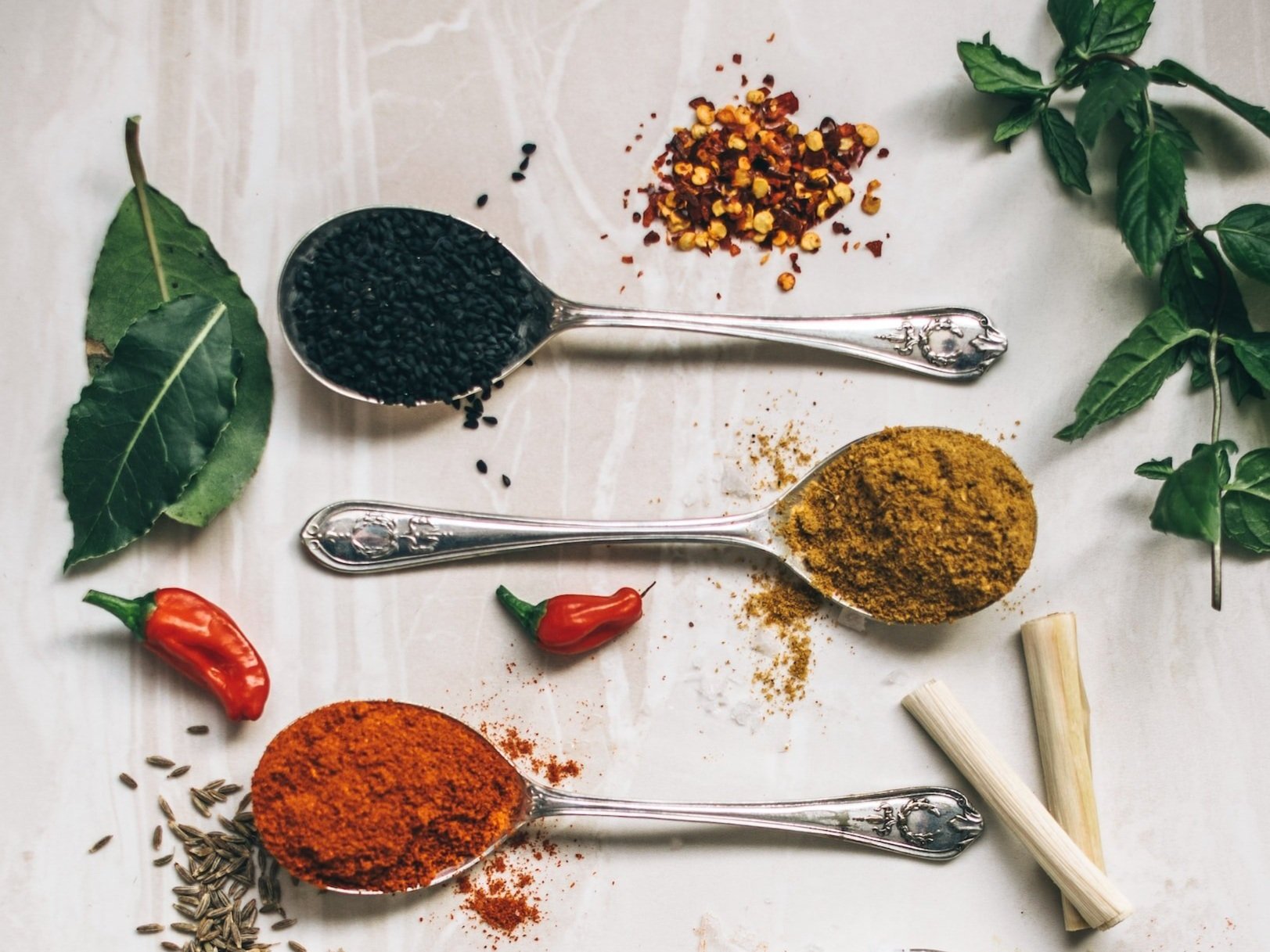 unsplash-image-vA1L1jRTM70 An assortment of spices displayed on silver spoons, including black sesame seeds, chili flakes, red chili peppers, cumin, and curry powder, alongside fresh basil and bay leaves on a marble surface.